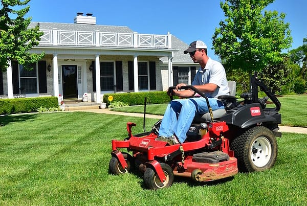 Person mowing a lawn with a riding lawn mower in front of a house.