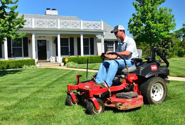 Person mowing a lawn with a riding lawn mower in front of a house.