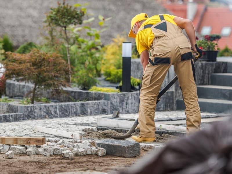 Worker laying stone pavers in a landscaped yard.